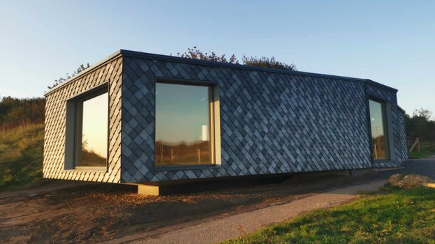 Corner view of the newly completed Whitburn Coastal Conservation Centre, a single story building clad in blue-grey diamond shaped tiles which look a little like fish scales. Low autumn sun hits three large windows, which reflect the blue sky and grass and shrubs of the surrounding clifftops.